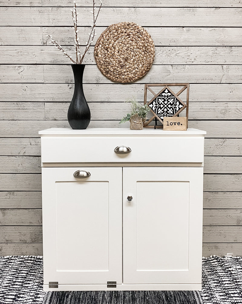 White tilt out trash bin with a storage cabinet and a drawer.  Handle options are gold, silver, or black. Panel options are cedar look panel, chalkboard, barn metal, or flat front.